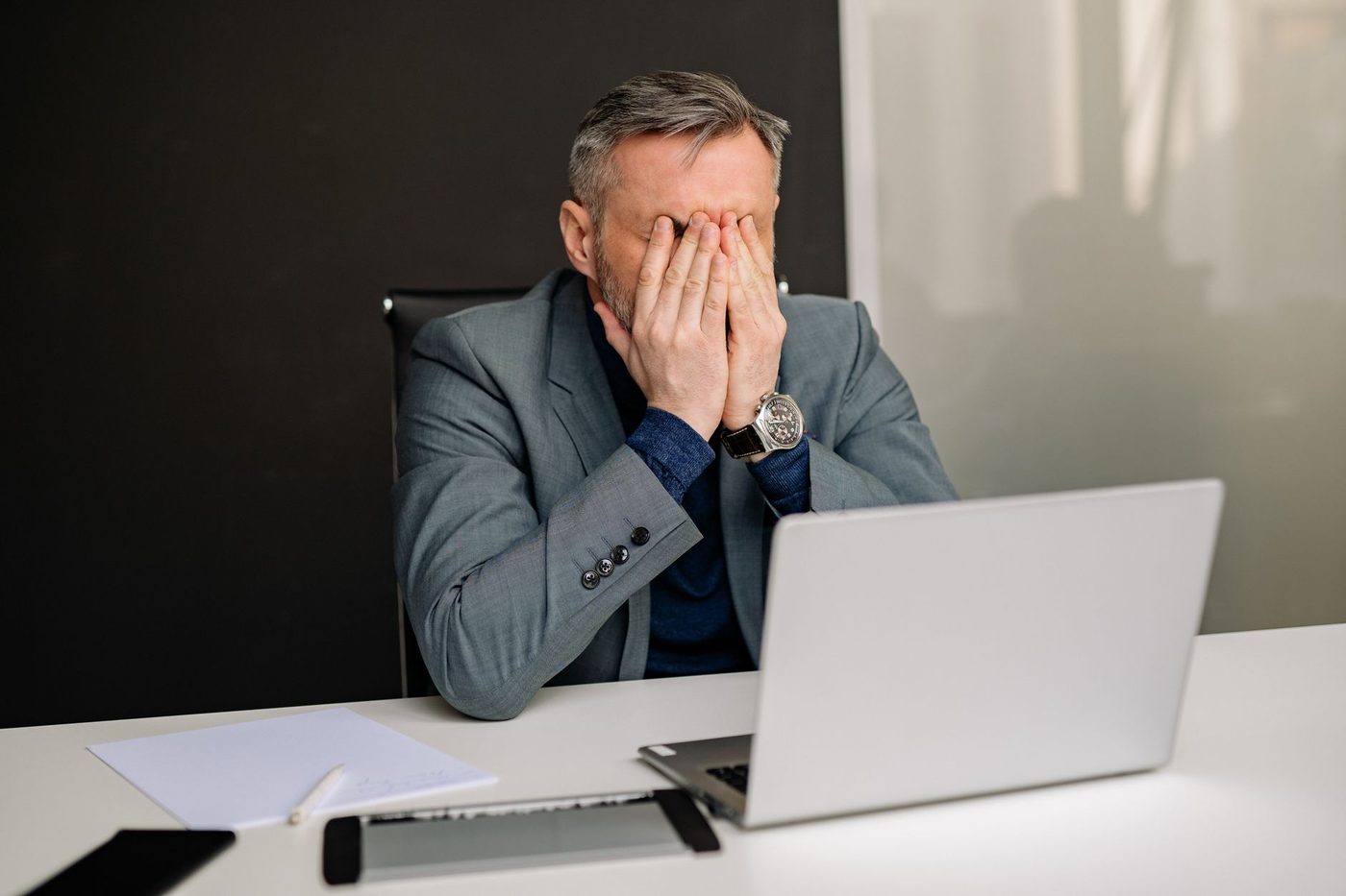 Homem de terno, sentado à mesa do escritório, com expressão de frustração diante do notebook.